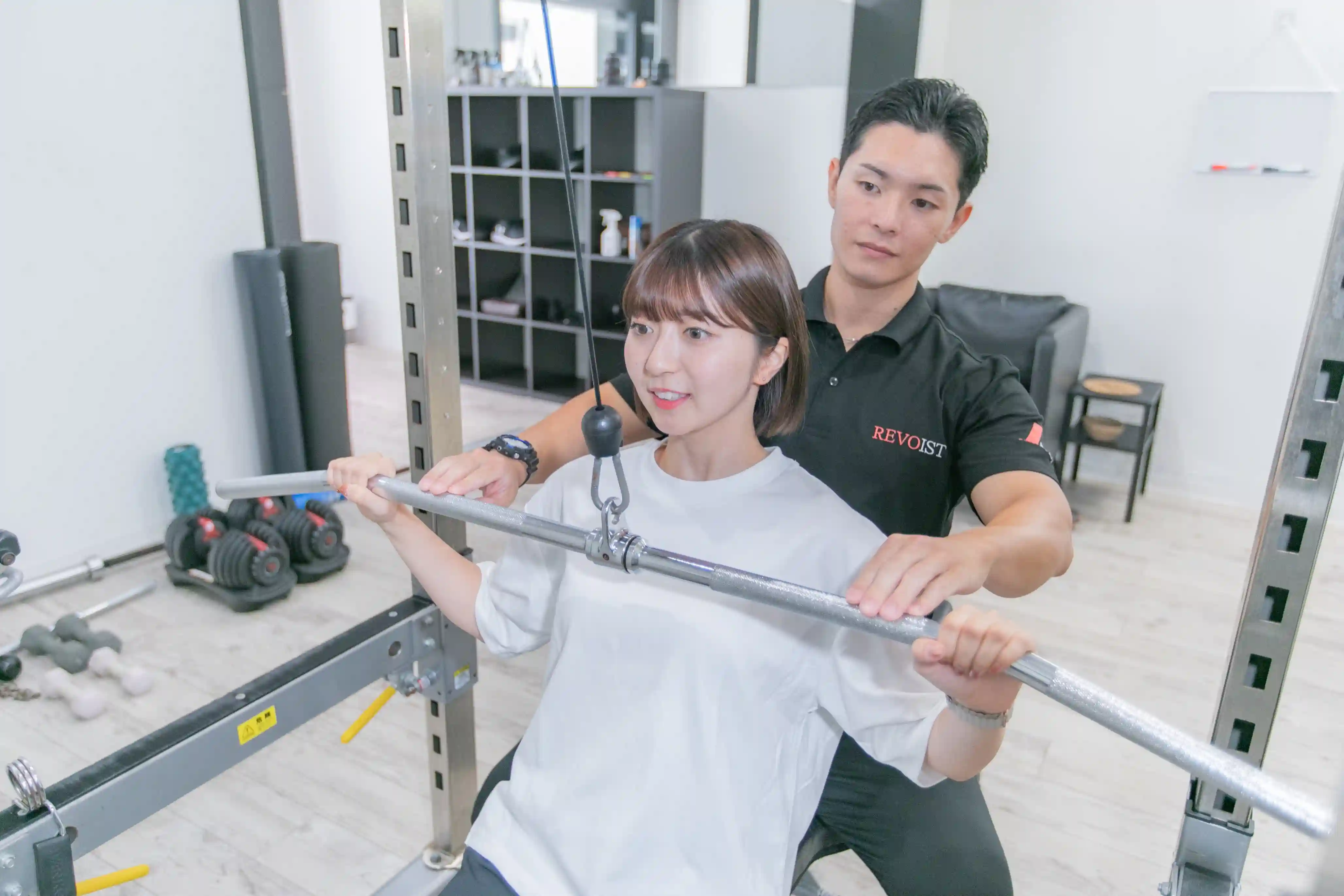 浦安のパーソナルジム「REVOIST」のトレーニングメニュー例。トレーナーの丁寧なフォーム指導を受けながら、背中を効果的に鍛えるラットプルダウンを行う女性。