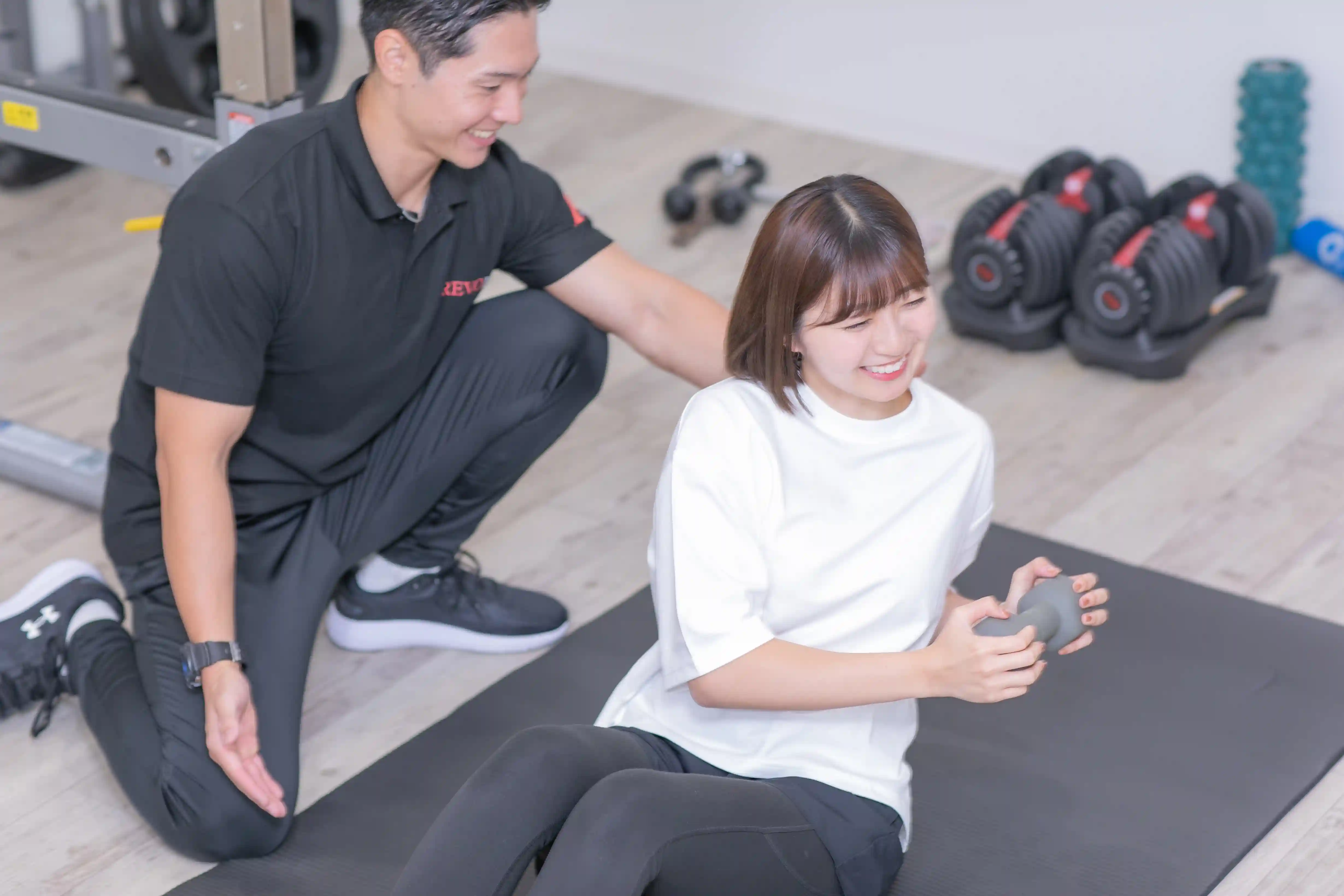 浦安のパーソナルジム「REVOIST」のトレーニングメニュー例。トレーナーの丁寧なサポートを受けながら、体幹をしっかり鍛えるツイストクランチを笑顔で行う女性。