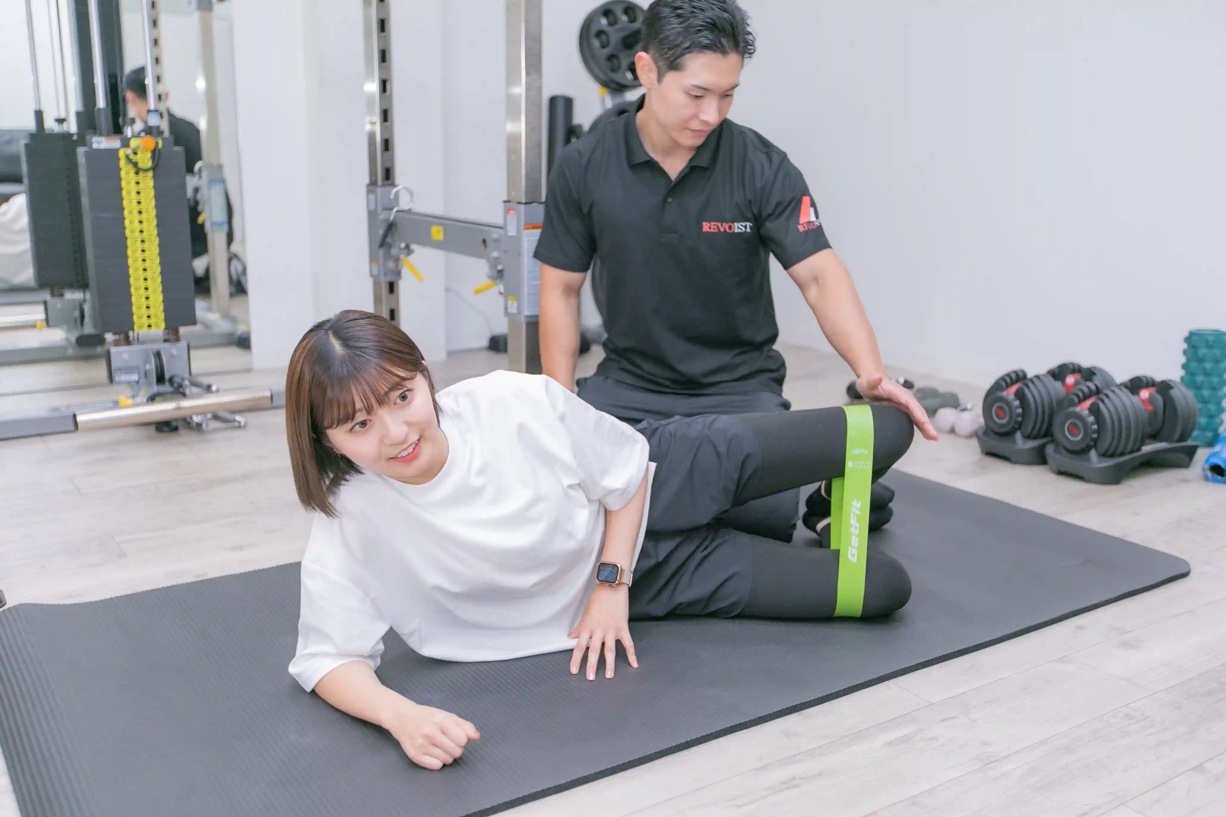 浦安のパーソナルジム「REVOIST」のトレーニングメニュー例。トレーナーのサポートを受けながら、ゴムバンドを使ってお尻を効果的に鍛えるクラムシェルを行う女性。