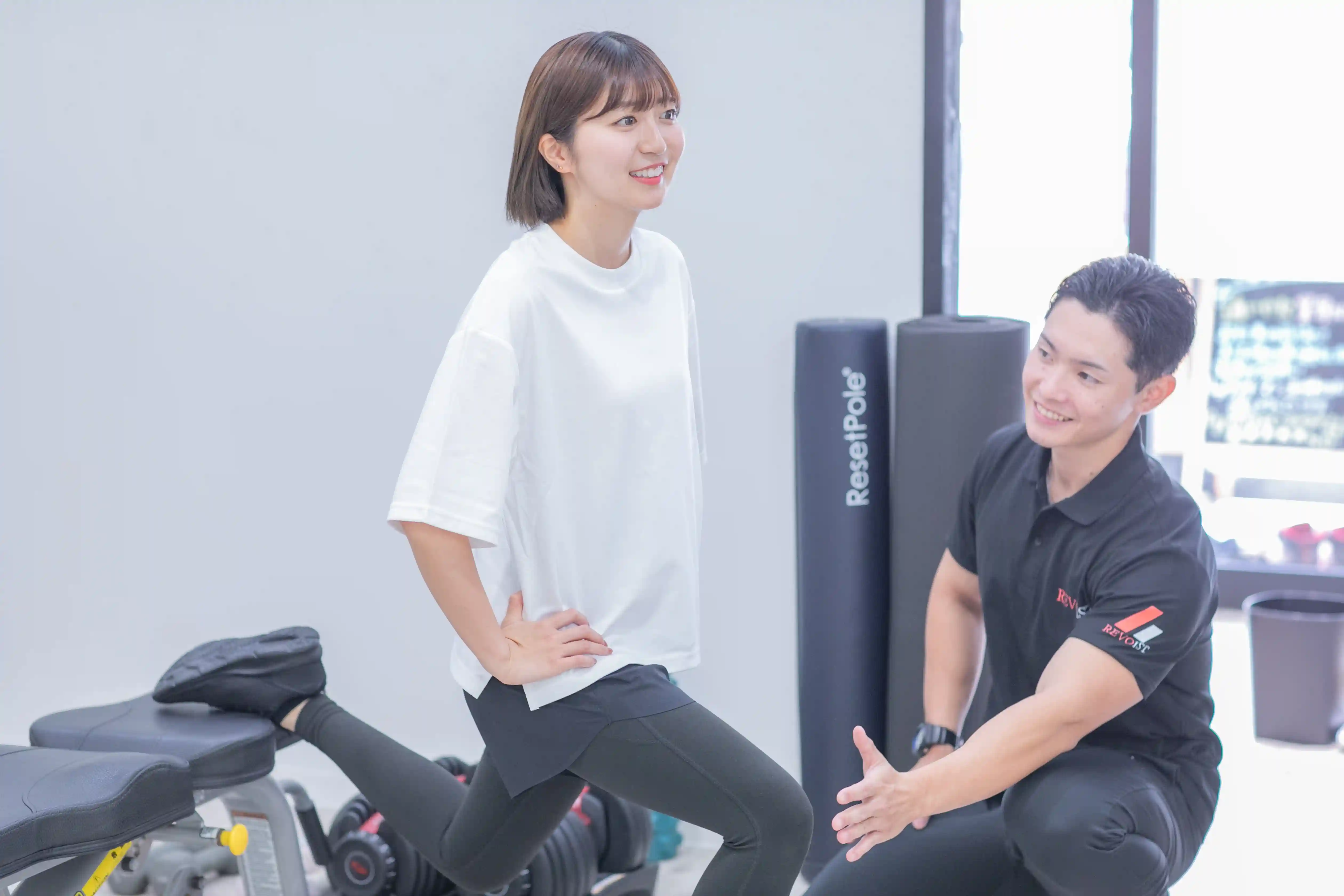 浦安のパーソナルジム「REVOIST」のトレーニングメニュー例。トレーナーのサポートのもと、下半身を効果的に鍛えるブルガリアンスクワットを笑顔で行う女性。