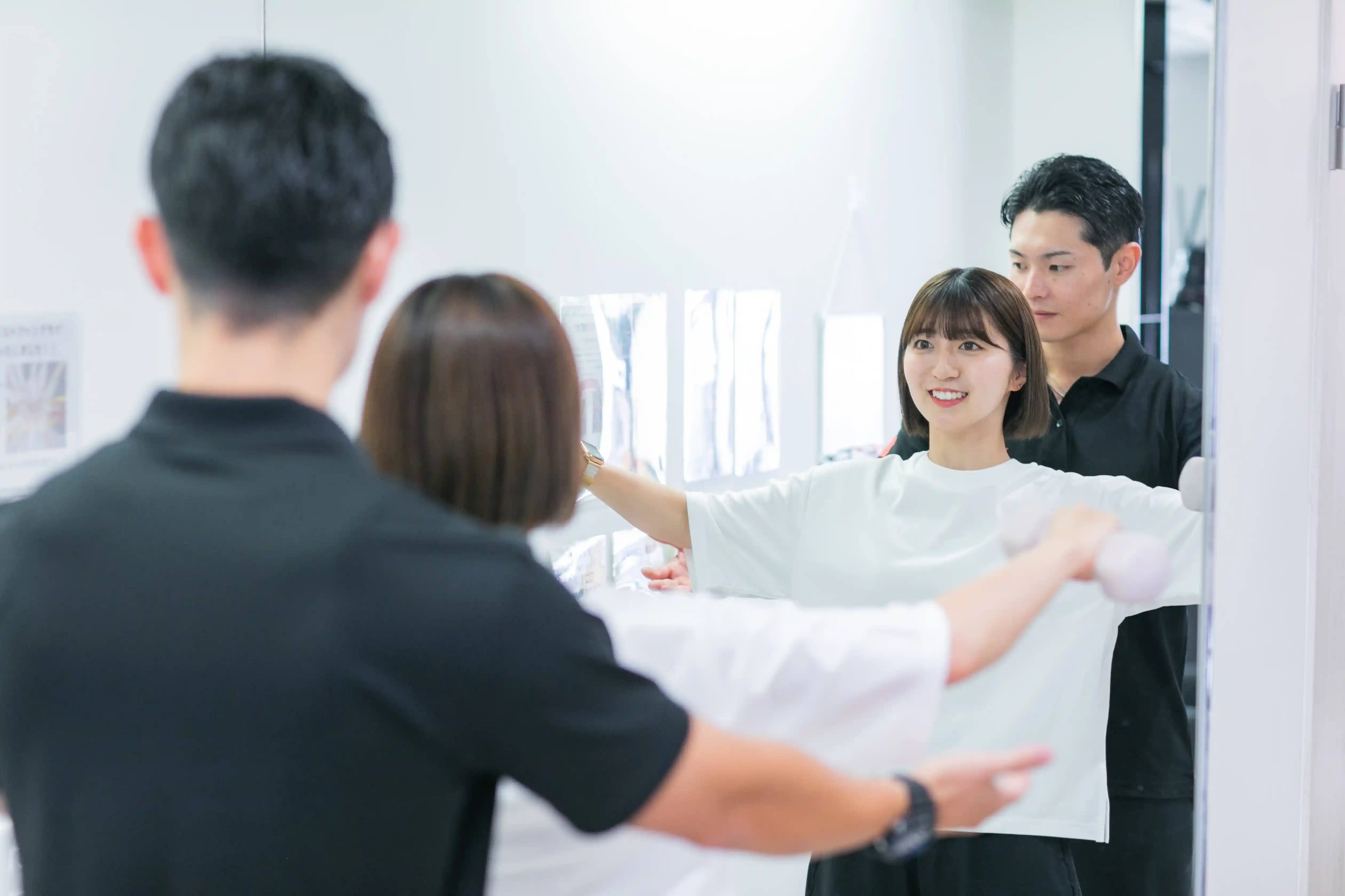 浦安のパーソナルジム「REVOIST」で、トレーナーと共に鏡でフォームを確認しながらダンベルサイドレイズ（ラテラルレイズ）を行う女性。週1回のオーダーメイドメニューで効果的なトレーニングをサポートします。