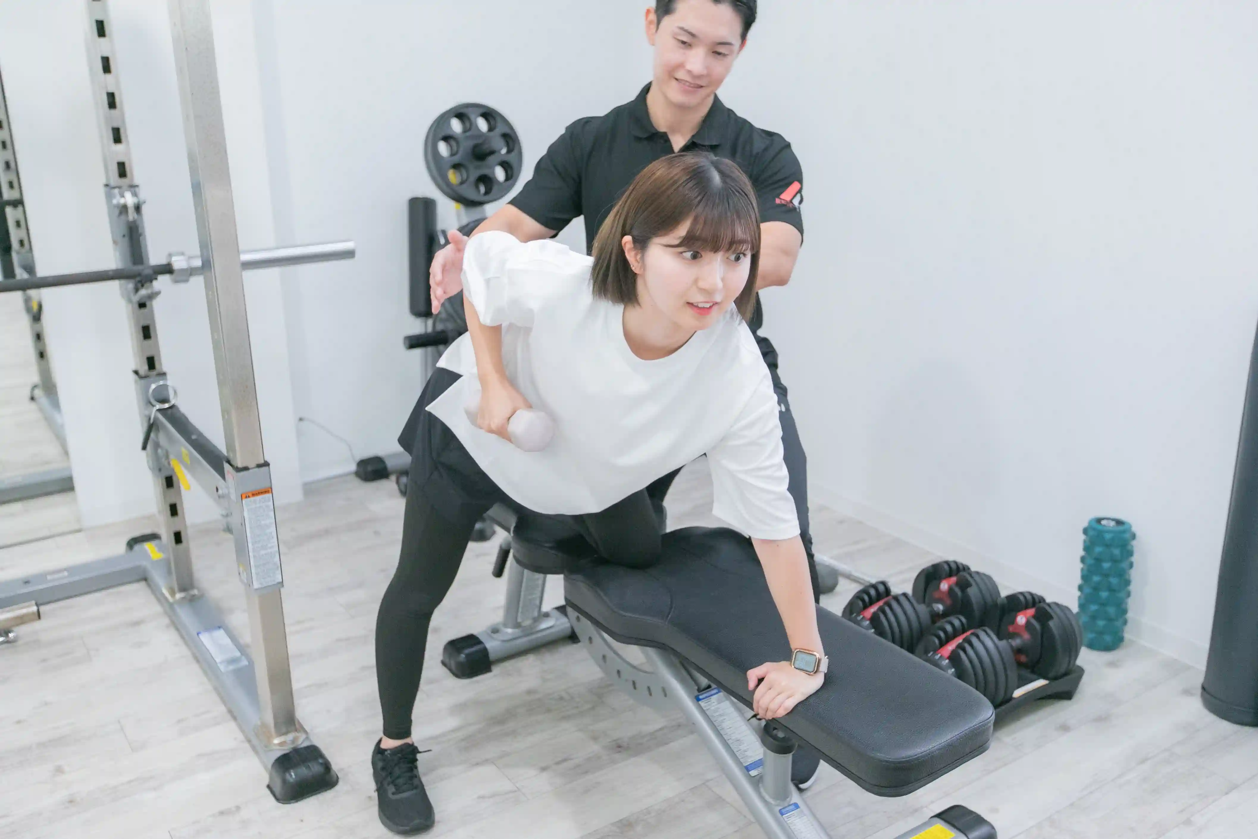 浦安のパーソナルジム「REVOIST」のトレーニングメニュー例。トレーナーのサポートを受けながら、二の腕を効果的に引き締めるダンベルキックバックを行う女性。